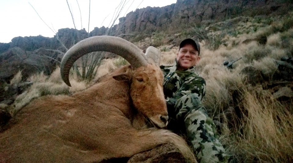 Hunter lying beside an aoudad ram in tall grass with cliffs rising behind