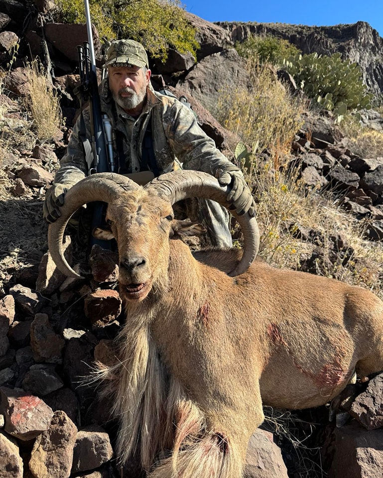 Hunter standing on a rocky slope holding an aoudad ram upright by the horns, rifle slung