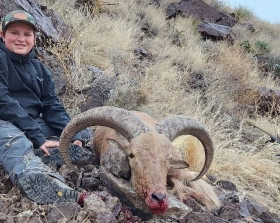 Young hunter sitting beside the heavy curled horns of an aoudad ram on a rocky slope